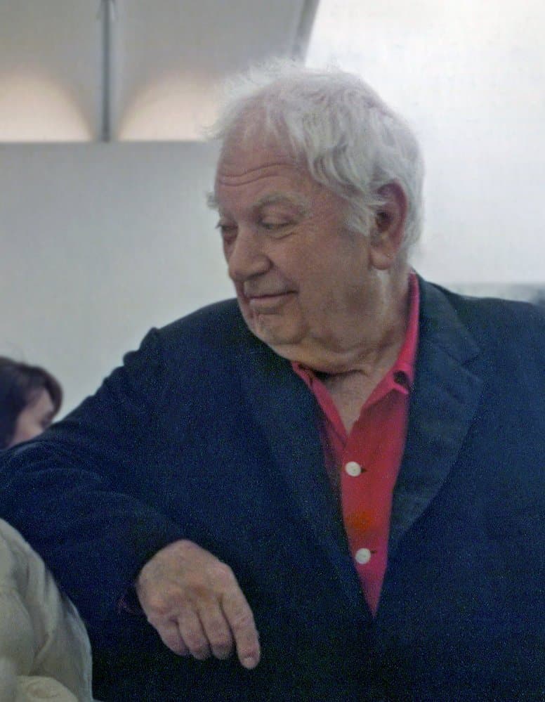 Alexander Calder, white-haired, in a blue jacket and red shirt, looking down.