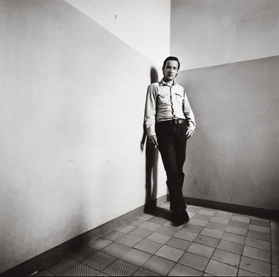 Blinky Palermo leaning against a white wall in the corner of a room.