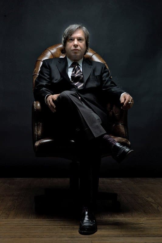 George Condo in a dark suit and striped tie, seated in a leather armchair.