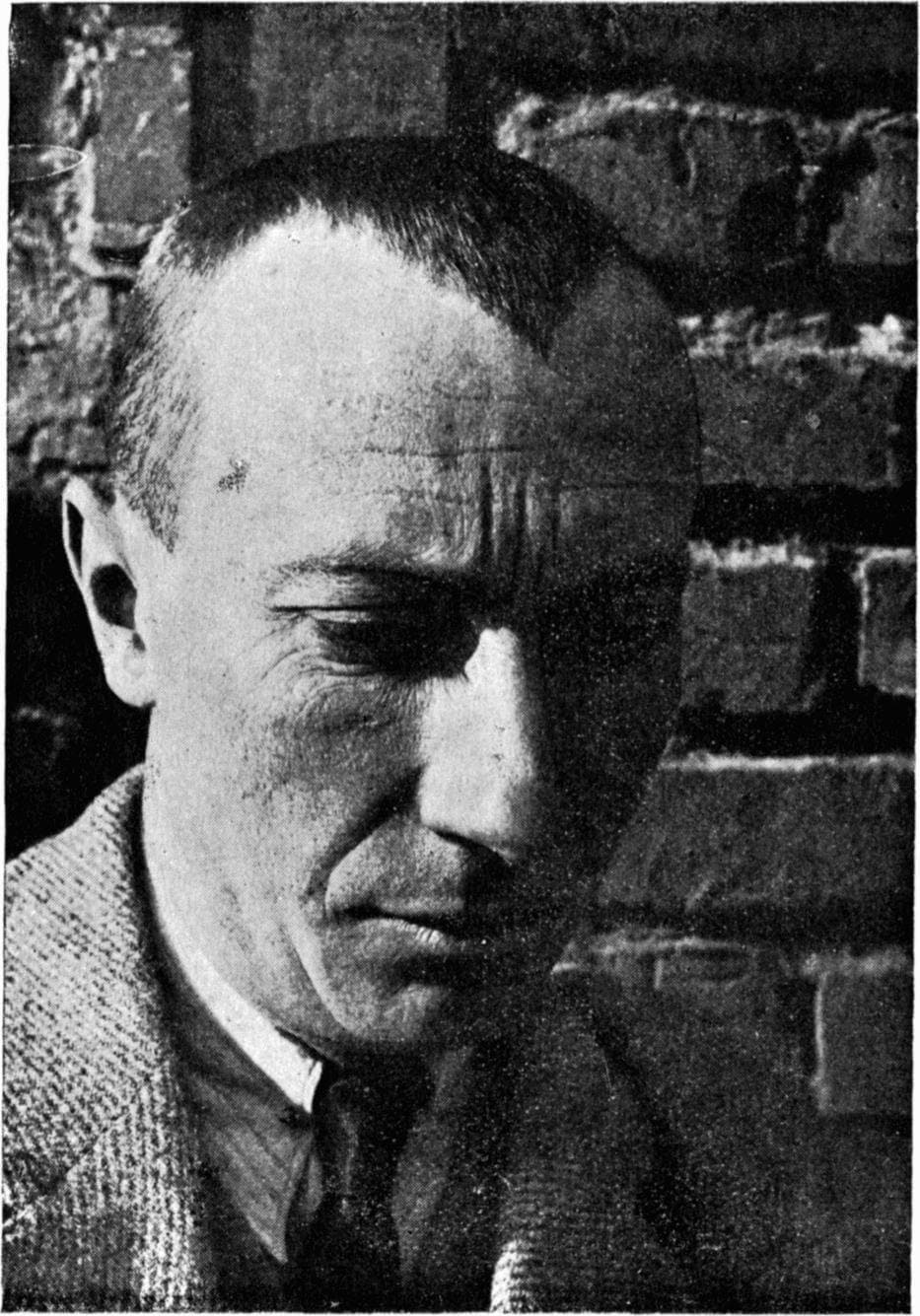 Jean Arp in half shadow, head bowed, against a brick wall.