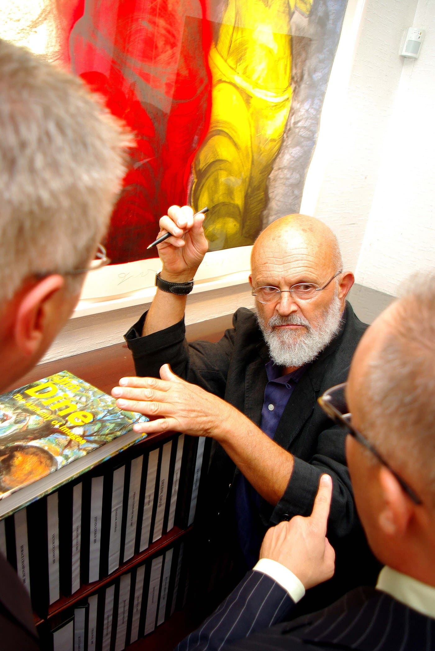 Jim Dine, bald and white-bearded, signing at a table among visitors to a gallery.