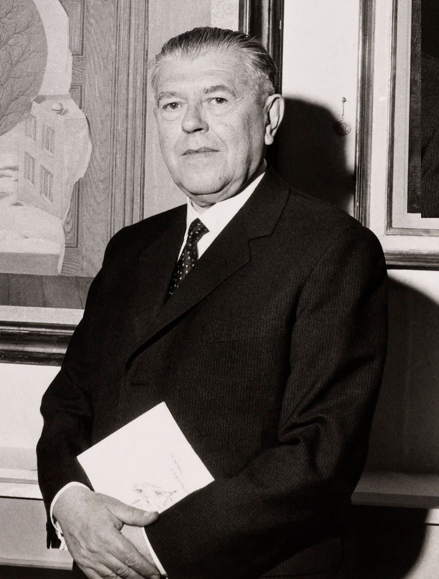 René Magritte in a dark suit and tie, in front of one of his paintings.