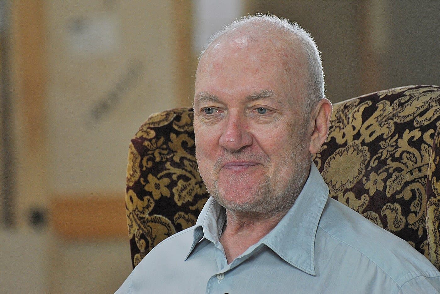 Sean Scully in a pale blue shirt, seated in a patterned armchair in his studio.