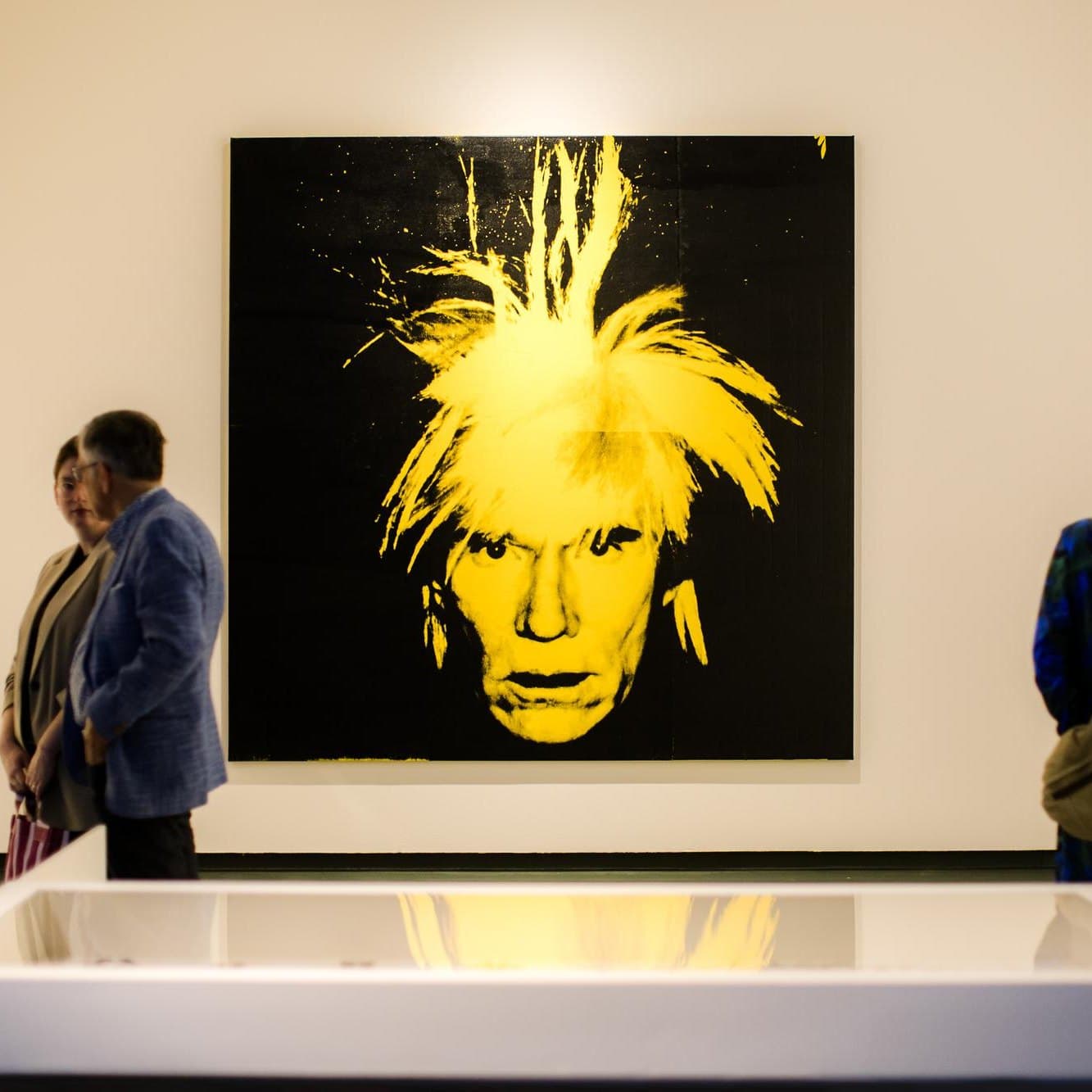 Visitors standing either side of Warhol's yellow-on-black fright-wig self-portrait hung on a gallery wall, with a vitrine in the foreground