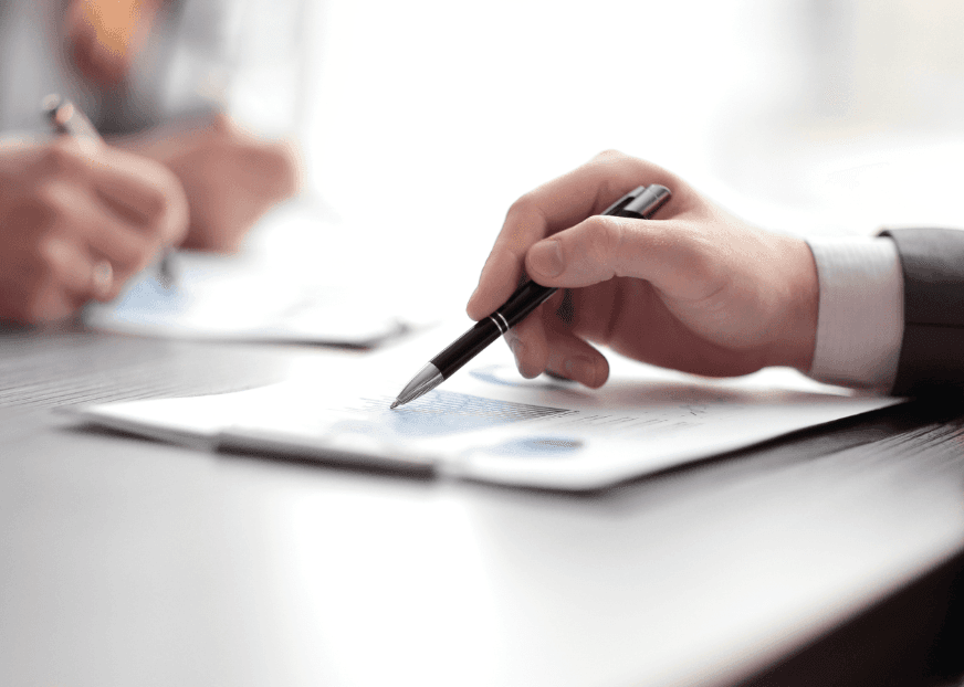 A hand resting the tip of a pen on a printed document of figures and charts on a desk, with a second person writing on their own copy behind
