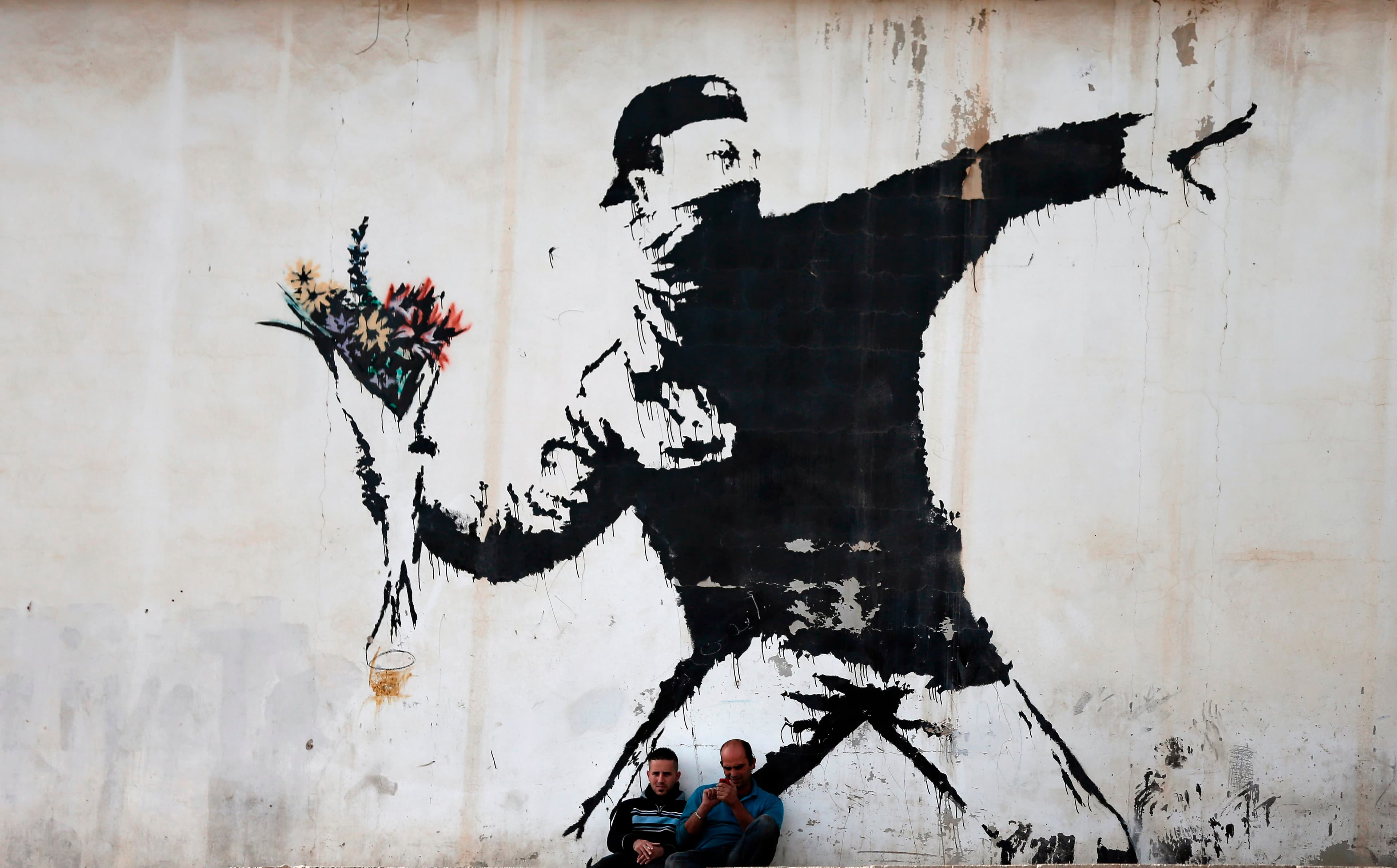 Banksy's Flower Thrower stencilled on a wall in the West Bank: a masked figure winding up to throw, holding a bouquet of flowers instead of a missile, with two men sitting on the ground beneath it