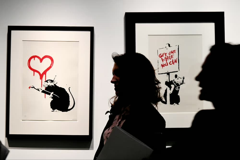 Two framed Banksy rat screenprints hanging on a gallery wall — Love Rat and Get Out While You Can — with visitors silhouetted in the foreground