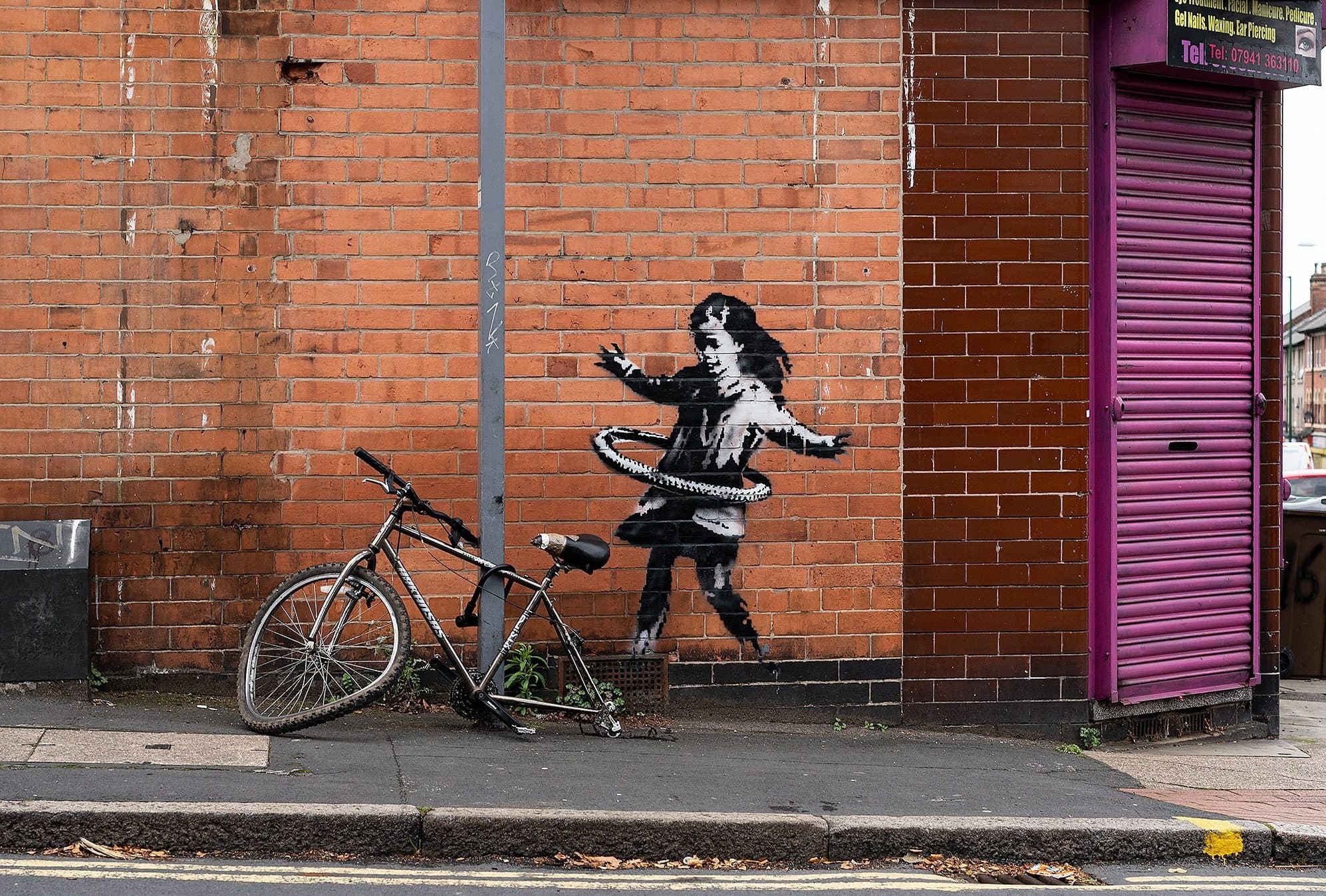 A Banksy stencil of a girl hula-hooping with a bicycle wheel, sprayed on a red brick corner beside a purple shop shutter, with a real bicycle missing its back wheel chained to the lamppost in front of it
