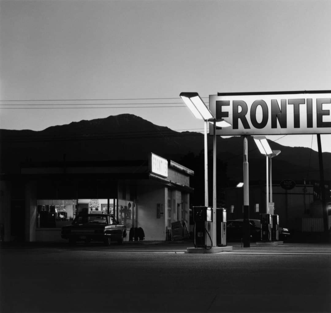 Robert Adams black and white photograph of a lit gas station at dusk below a mountain ridge