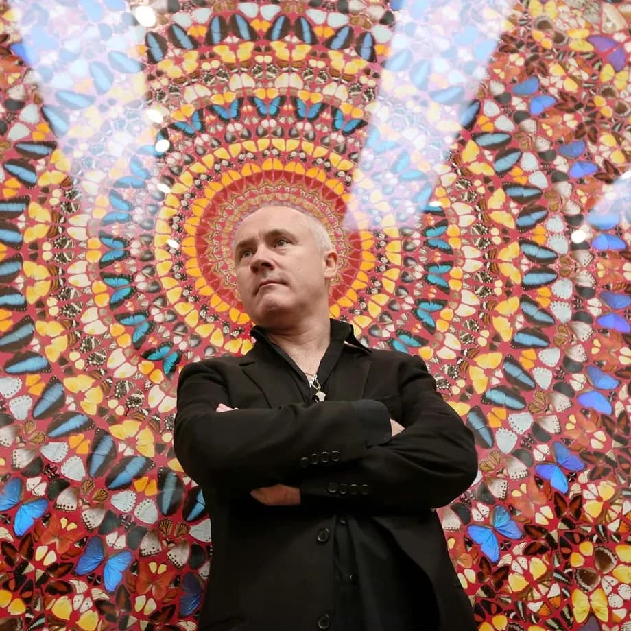 Damien Hirst standing with his arms folded in front of one of his butterfly kaleidoscope paintings, a dense circular mandala of coloured butterfly wings on a deep red ground