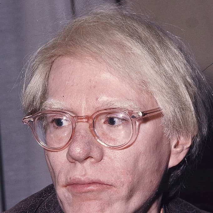 Andy Warhol photographed in 1975, close up in pale-framed glasses with his white hair swept across his forehead