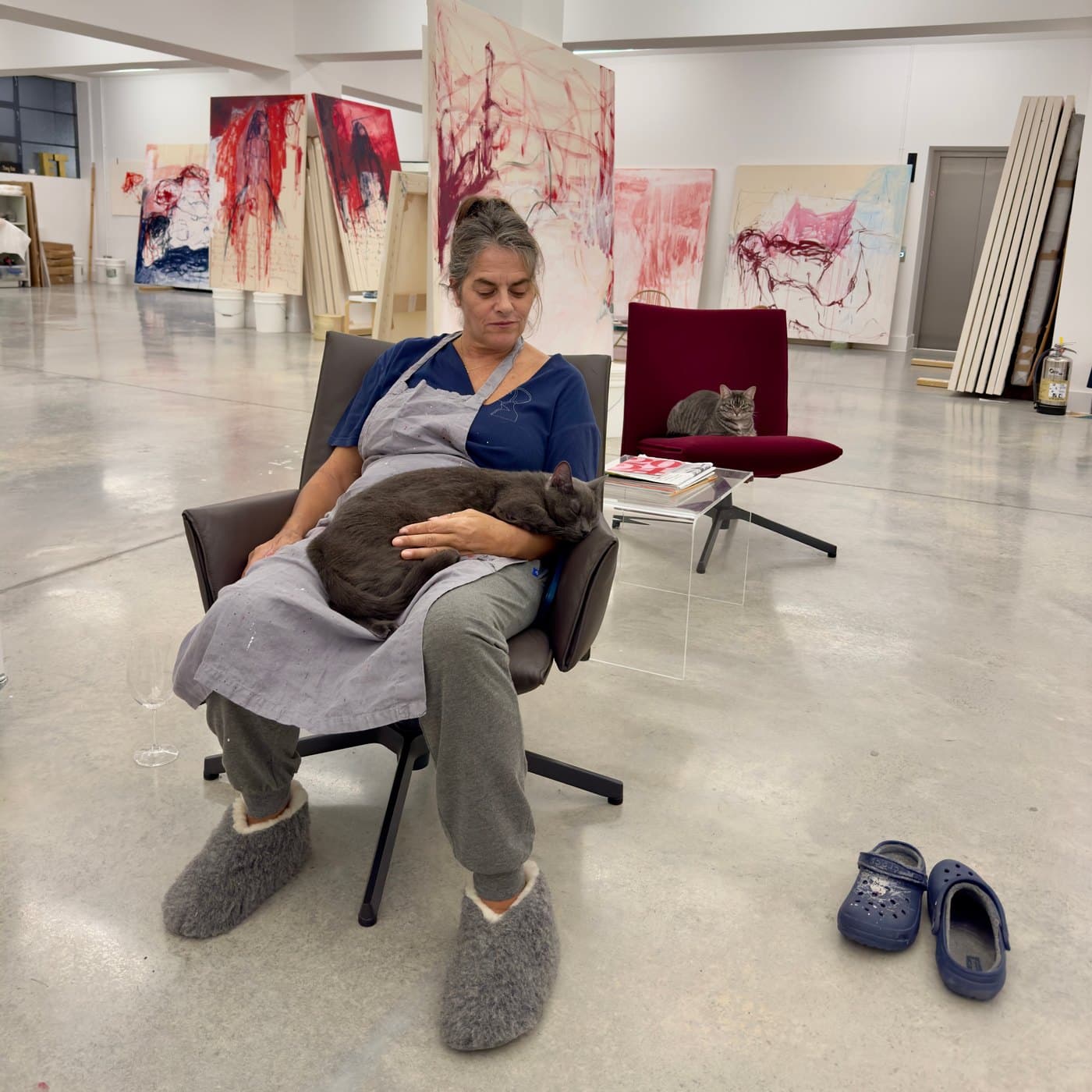 Tracey Emin seated in her studio with a cat asleep on her lap and a second cat on a chair behind, in front of a row of large red and pink paintings
