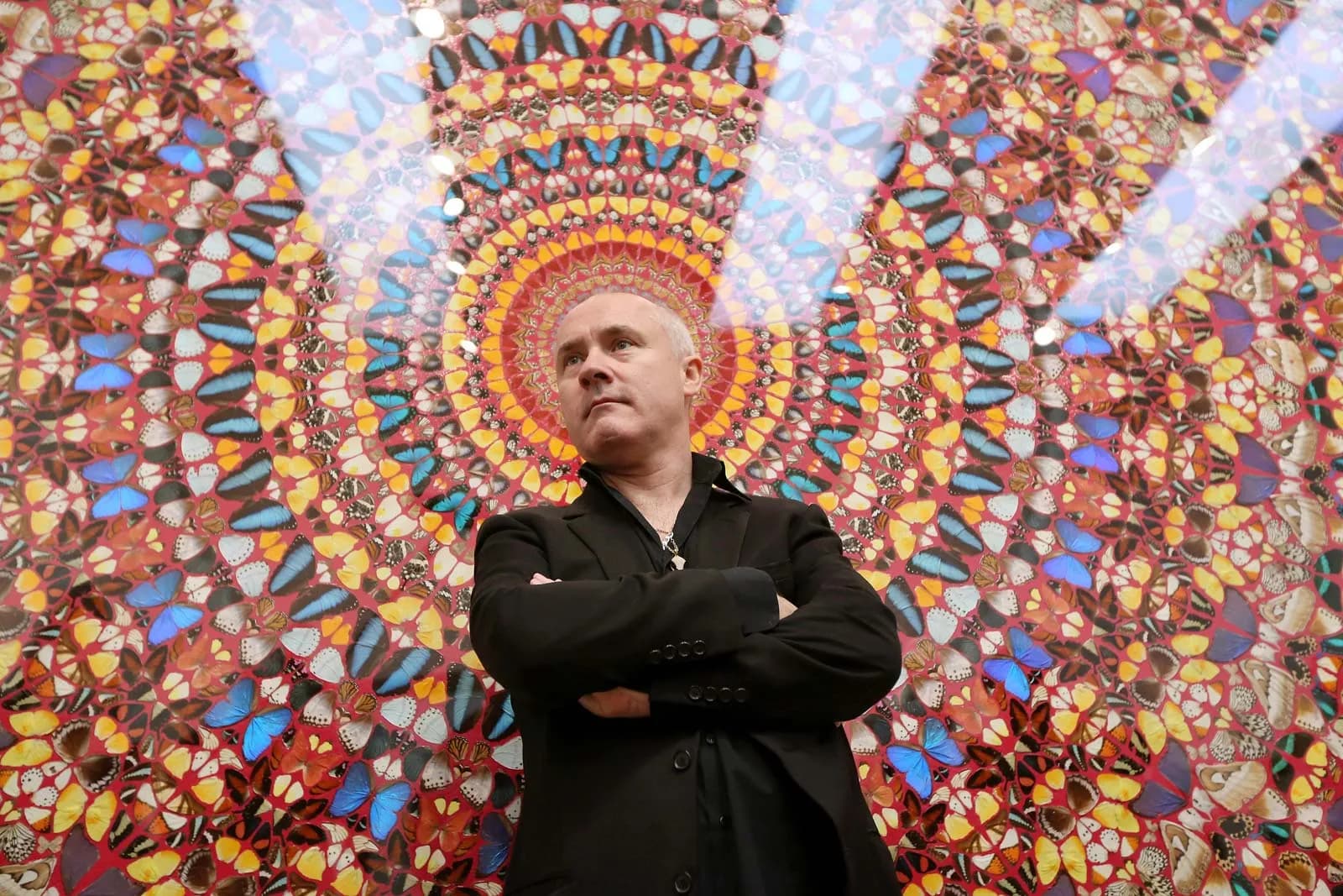 Damien Hirst standing with his arms folded in front of one of his butterfly kaleidoscope paintings, a dense circular mandala of coloured butterfly wings on a deep red ground