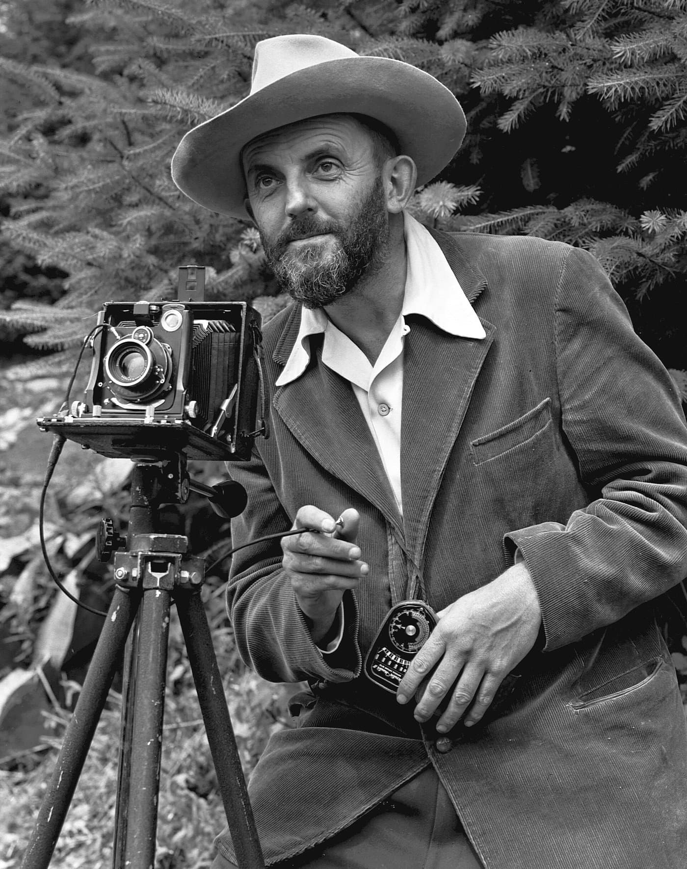 Ansel Adams in a wide-brimmed hat beside a large-format view camera on a tripod.