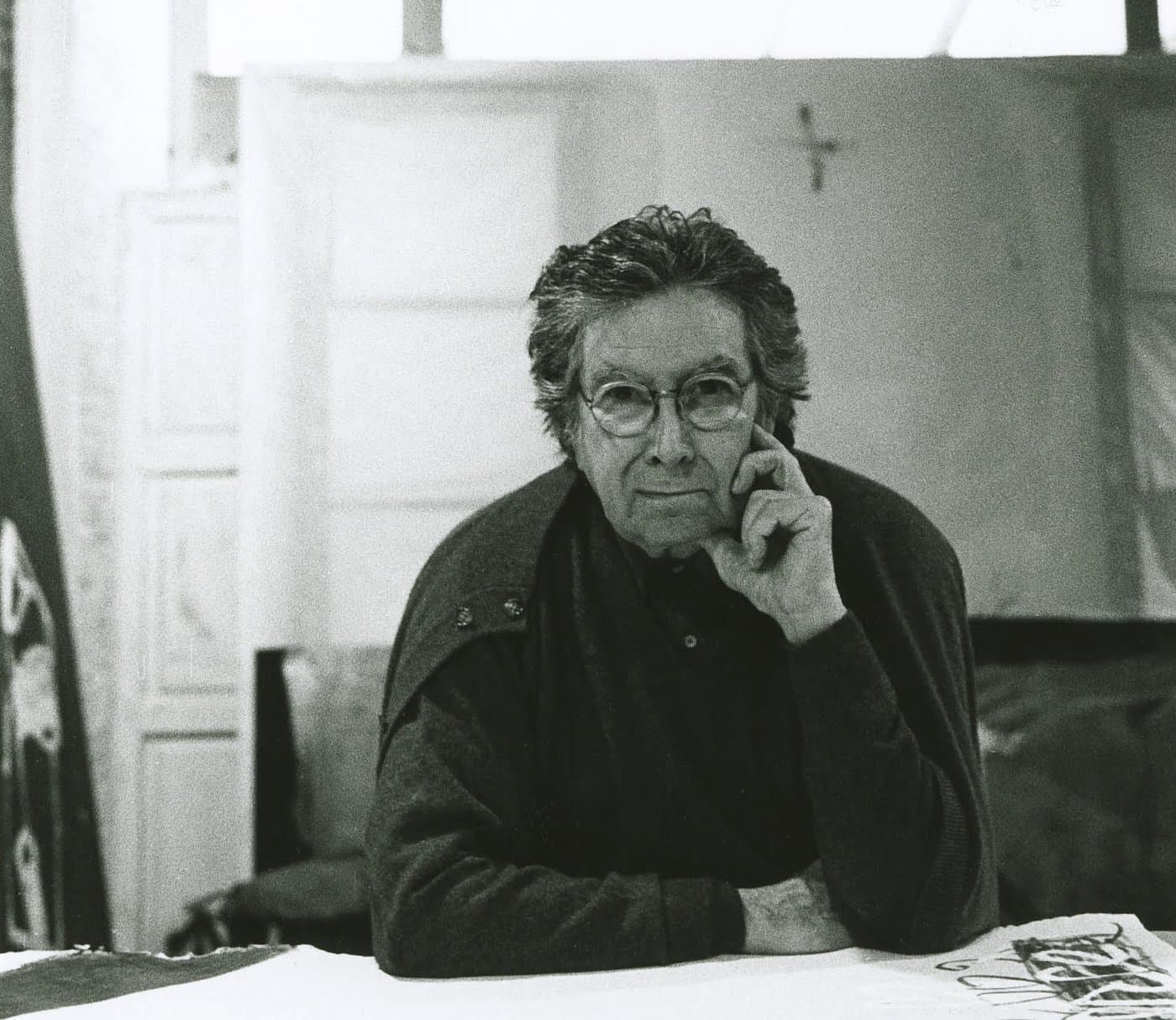 Antoni Tàpies in glasses, seated in his studio, a hand raised to his face.