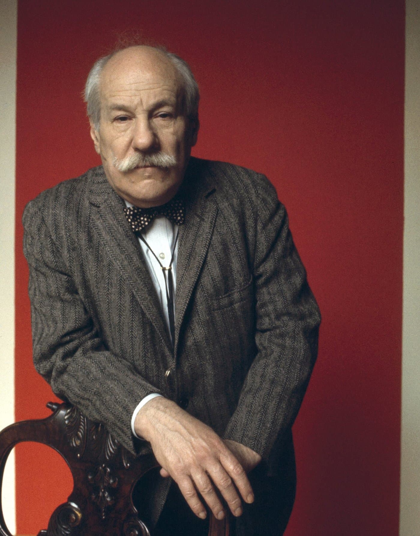 Barnett Newman with a white moustache and a bow tie, against a red ground.