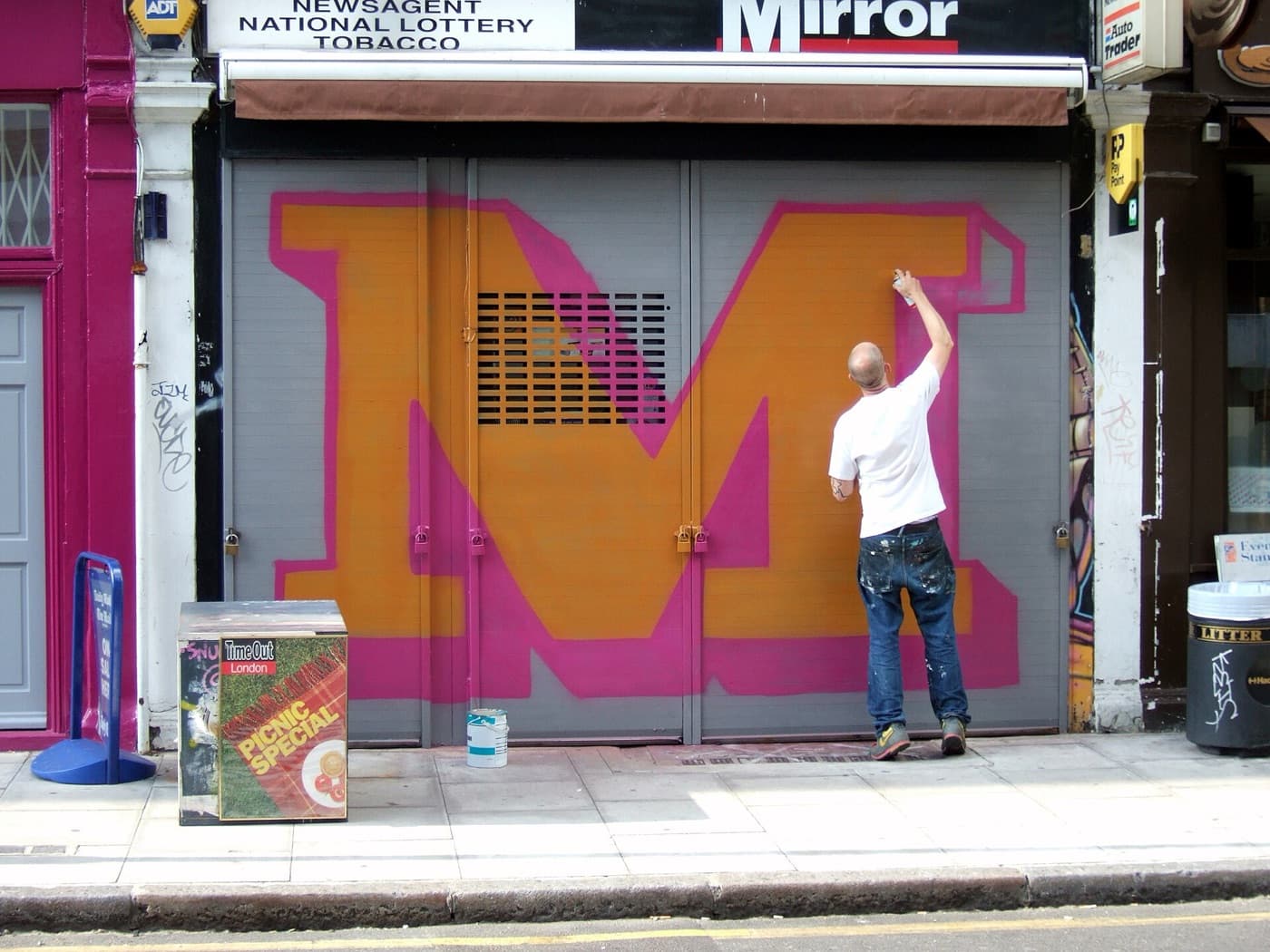 Ben Eine painting a large orange letter M on a pink shop shutter.