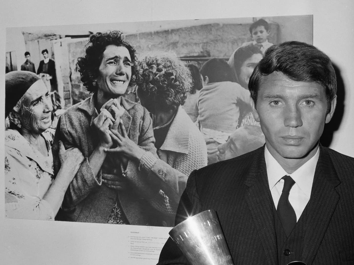 A young Don McCullin in a dark suit and tie beside an enlargement of his prizewinning photograph.