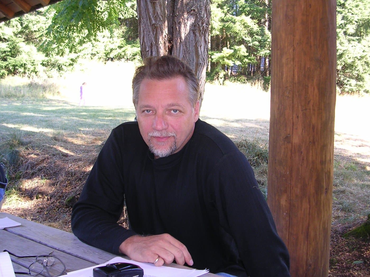 Edward Burtynsky in a black sweater, seated at a table outdoors under trees.