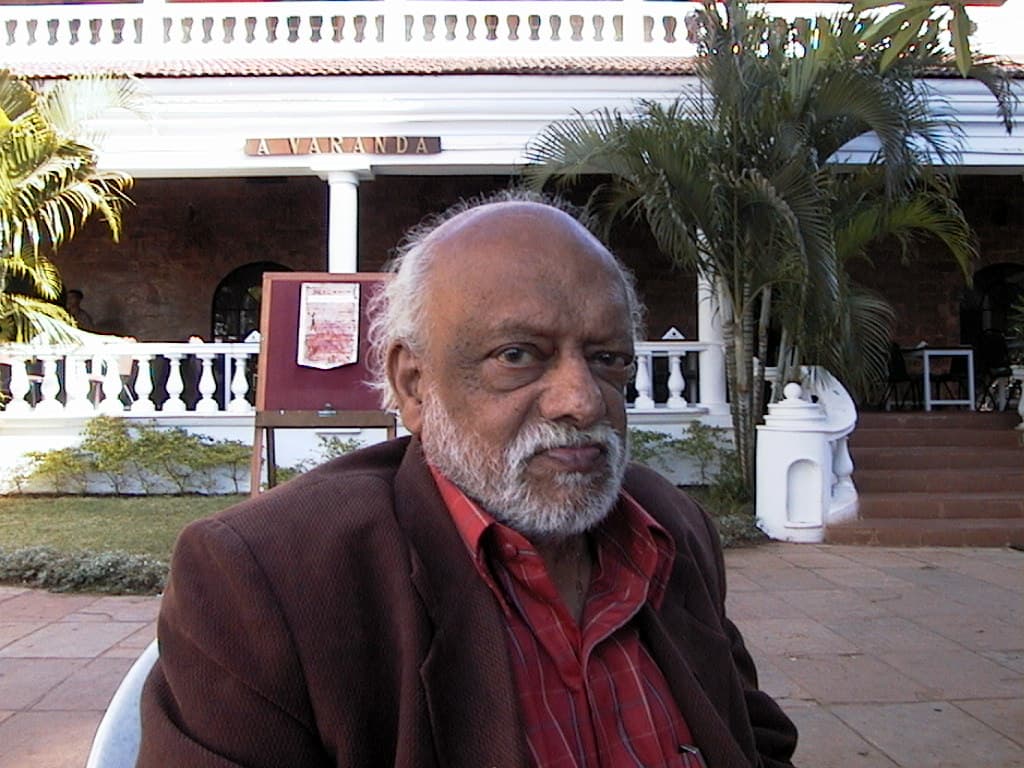 F. N. Souza, white-bearded, in a red shirt and dark jacket, seated outside a hotel.