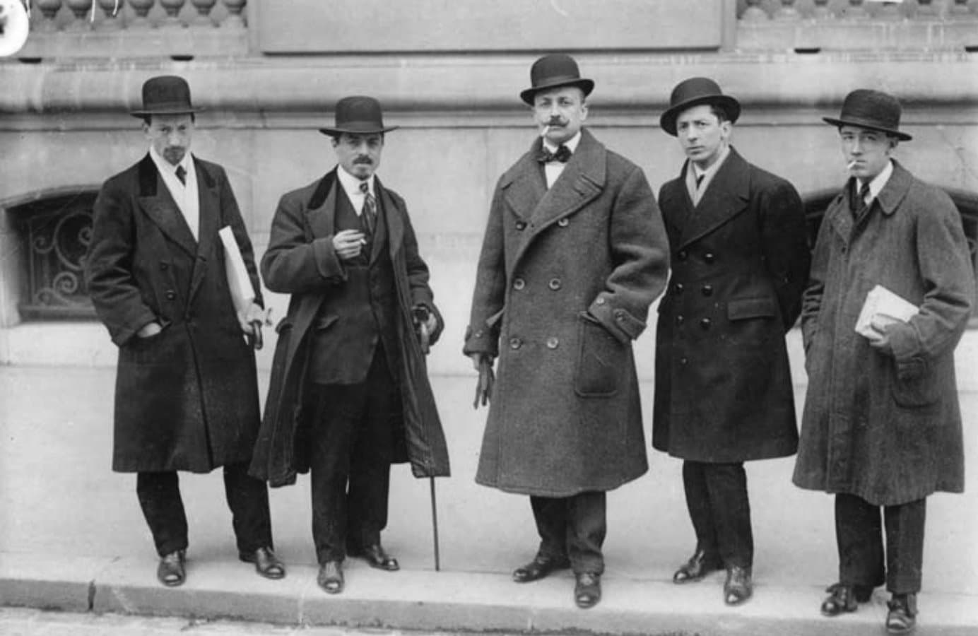 Five men in overcoats and bowler hats standing on a Paris pavement.