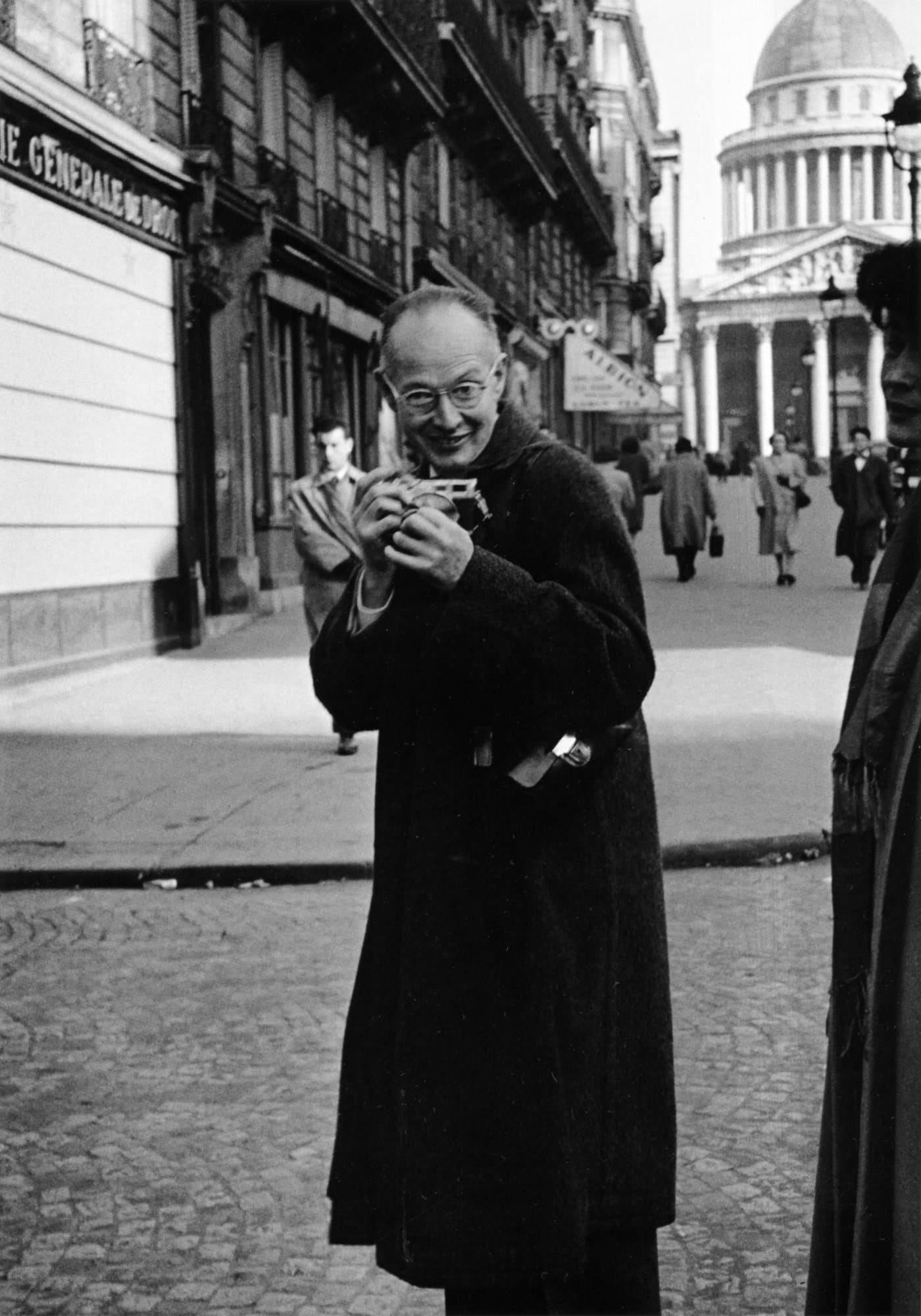 Henri Cartier-Bresson on a Paris street, holding a small camera to his chest.