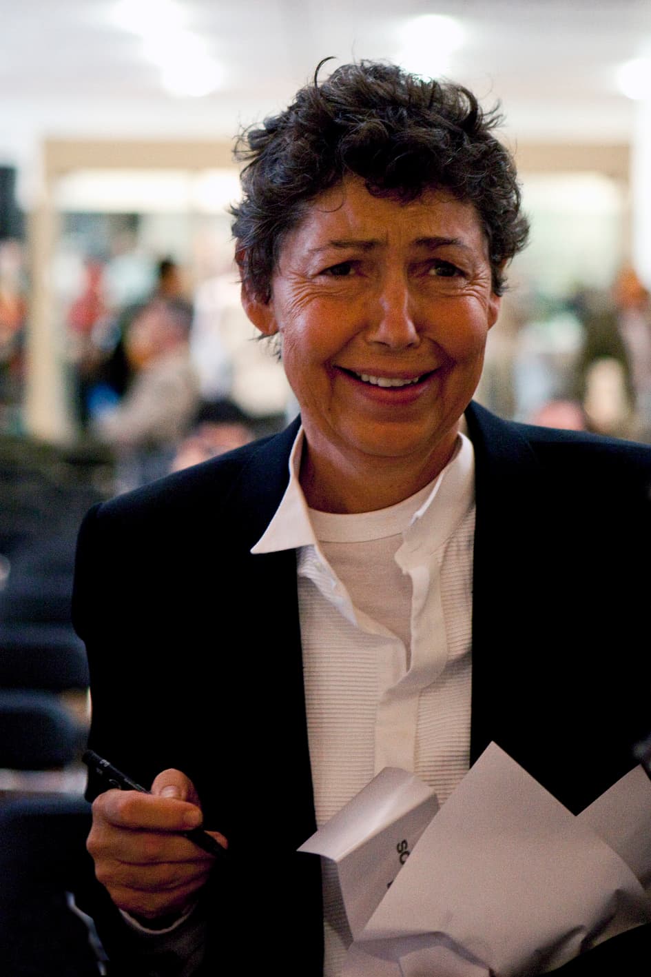 Isa Genzken, with short dark curly hair, smiling at an exhibition opening.