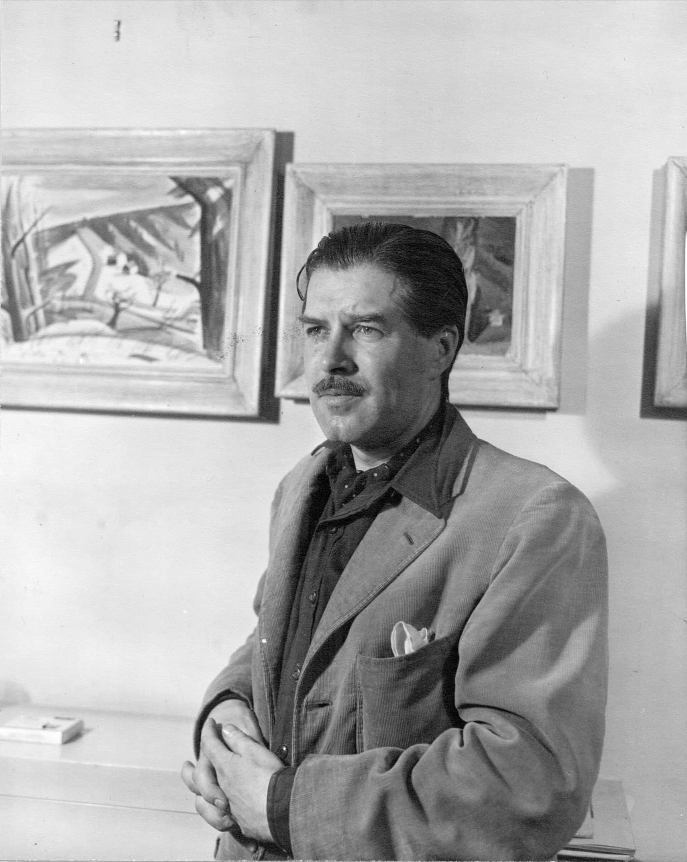 Jack Bush in a suit in front of framed paintings, in black and white.