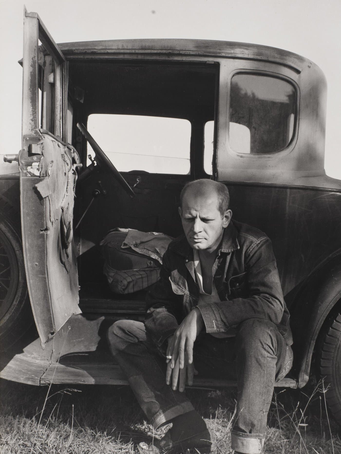 Jackson Pollock sitting on the running board of an old car, looking down.