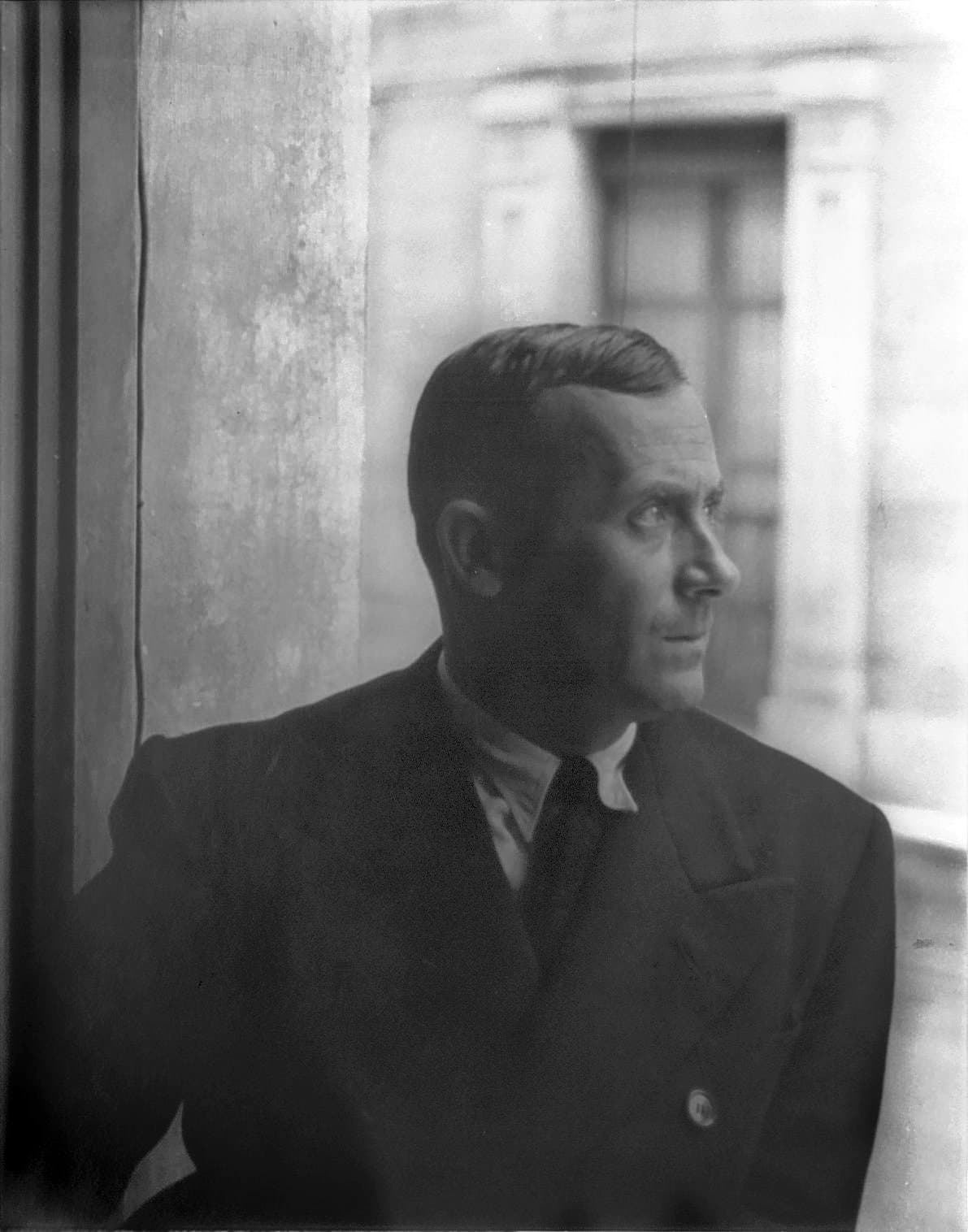 Joan Miró in profile, in a jacket and tie, beside a pale wall.