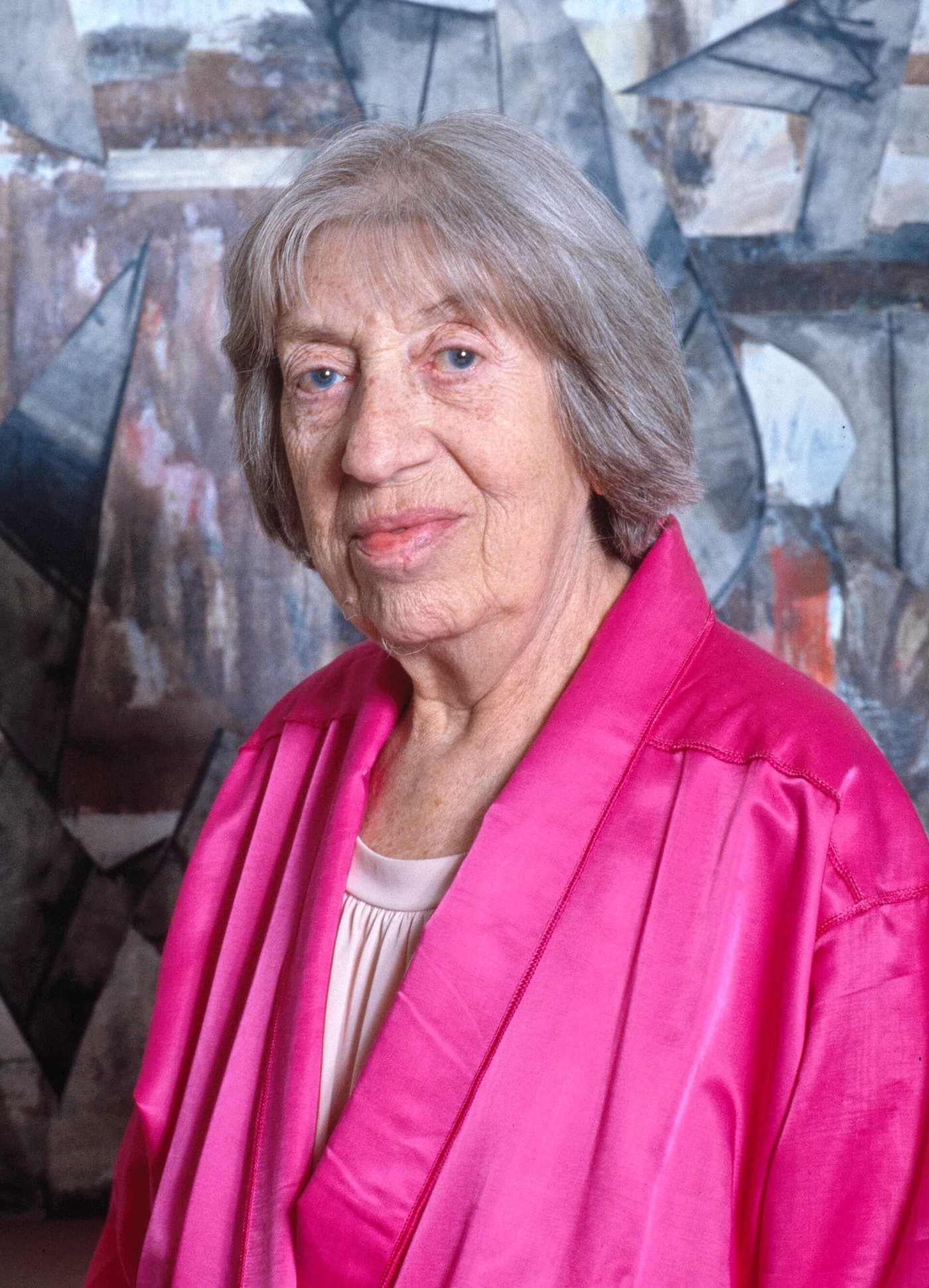 Lee Krasner with grey hair, in a pink jacket, in front of a painting.