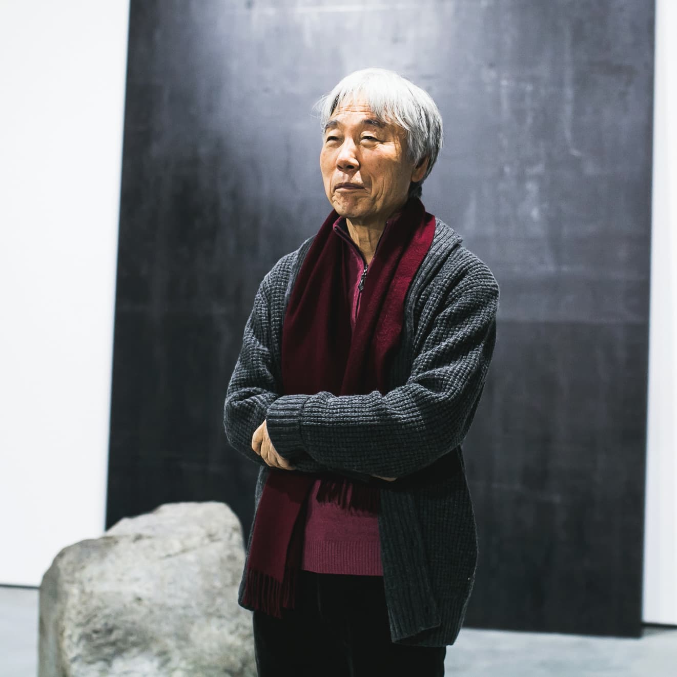 Lee Ufan, arms folded, in front of a large dark painting.