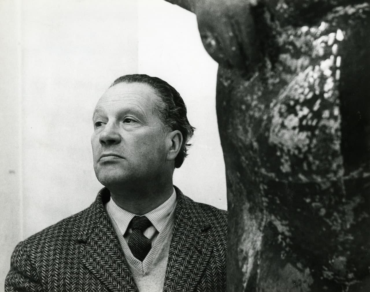 Marino Marini in a checked jacket beside a sculpture in his studio, in black and white.