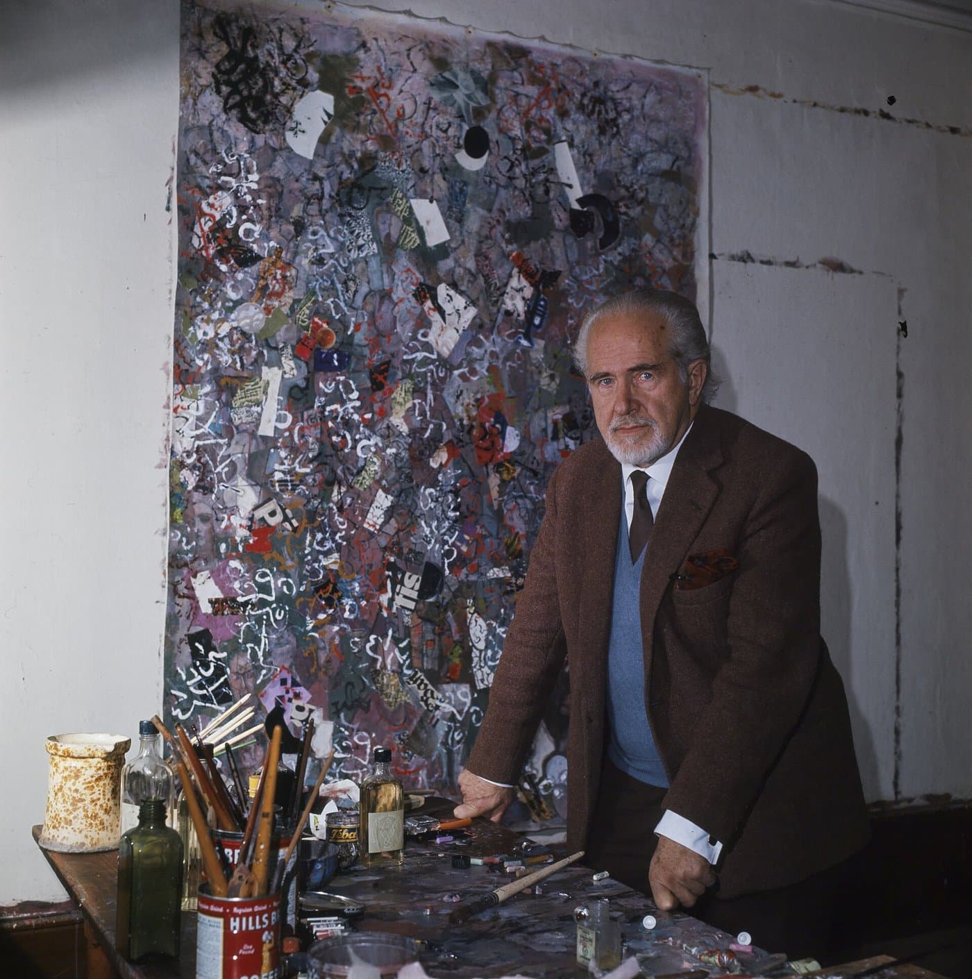 Mark Tobey, white-bearded, in a jacket and tie, seated in front of a large abstract painting.