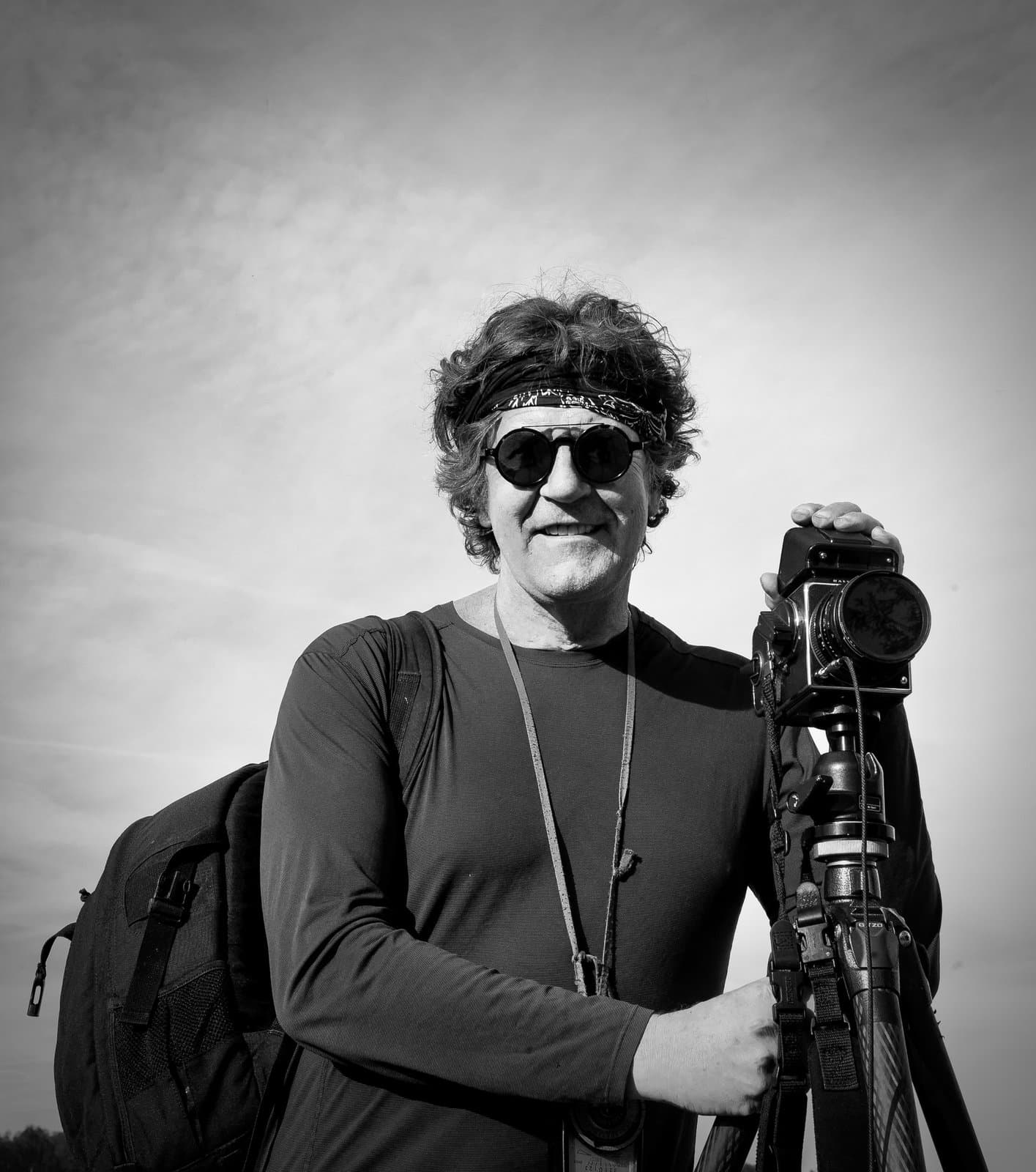 Michael Kenna in sunglasses and a headband beside a camera on a tripod, in black and white