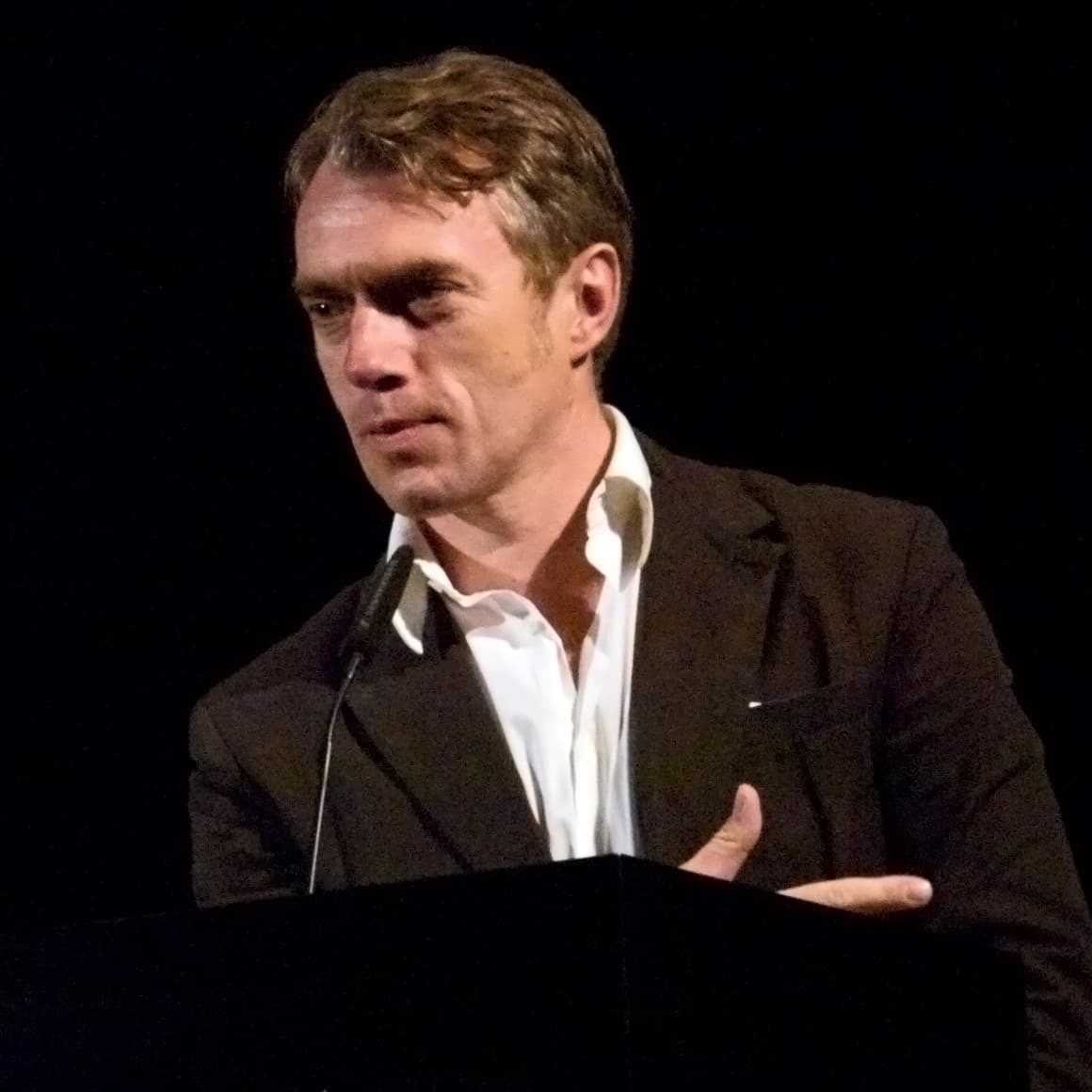 Neo Rauch in a dark jacket and open-necked white shirt at a lectern.