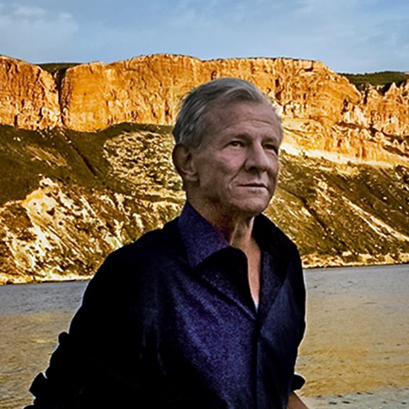 Peter Beard in a dark shirt, seated in front of cliffs above the sea.