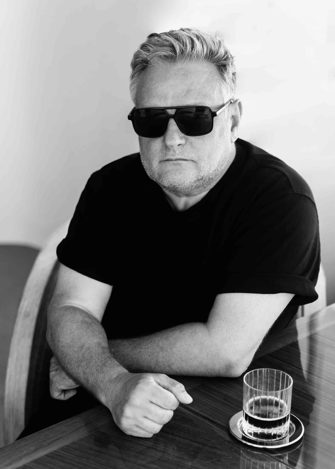 Rankin in dark glasses and a black T-shirt, seated at a table, in black and white