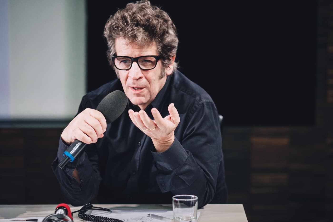 Robert Longo in black-rimmed glasses, speaking into a microphone.