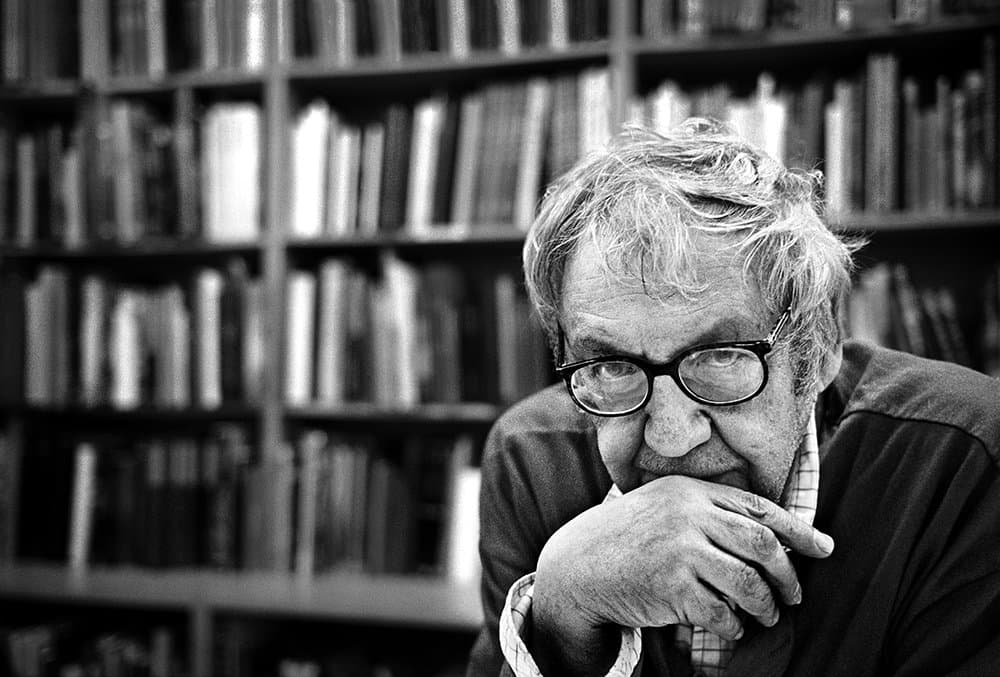 Saul Leiter in glasses, hand to his chin, in front of shelves of books