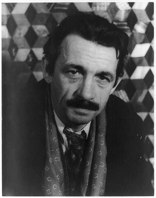 Thomas Hart Benton with dark hair and a thick moustache, in front of a patterned backdrop.