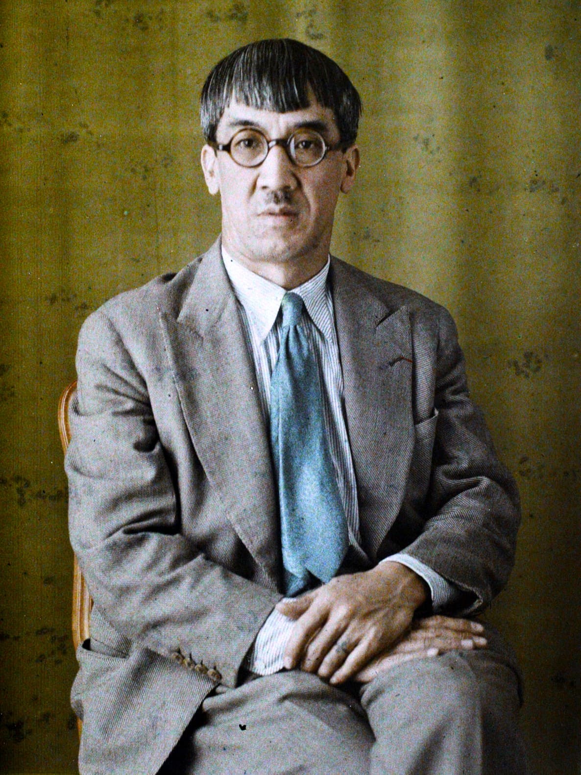 Foujita with his fringe and round glasses, in a grey suit and blue tie.