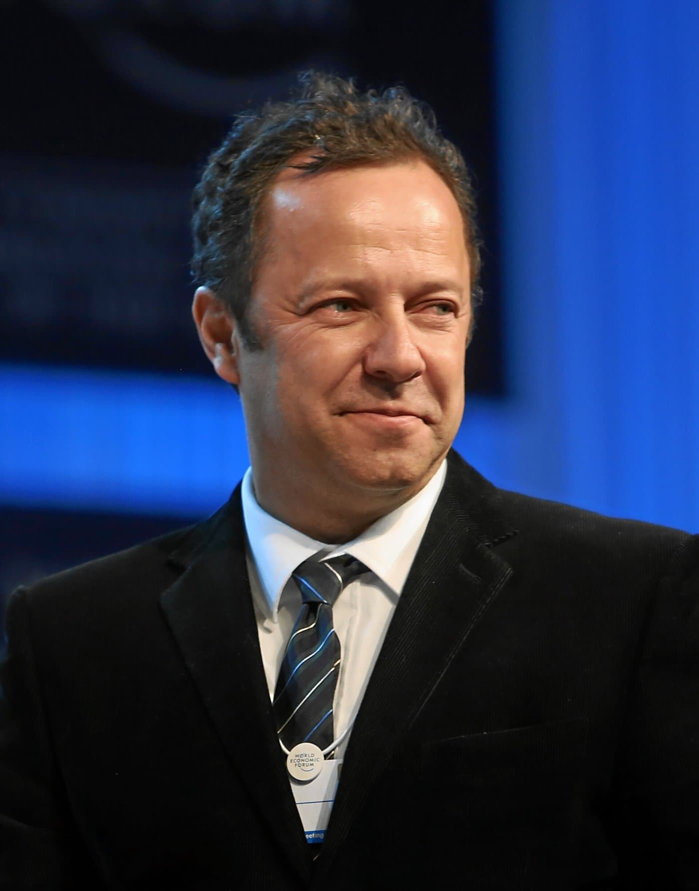 Vik Muniz in a dark suit and tie, smiling, against a blue background.