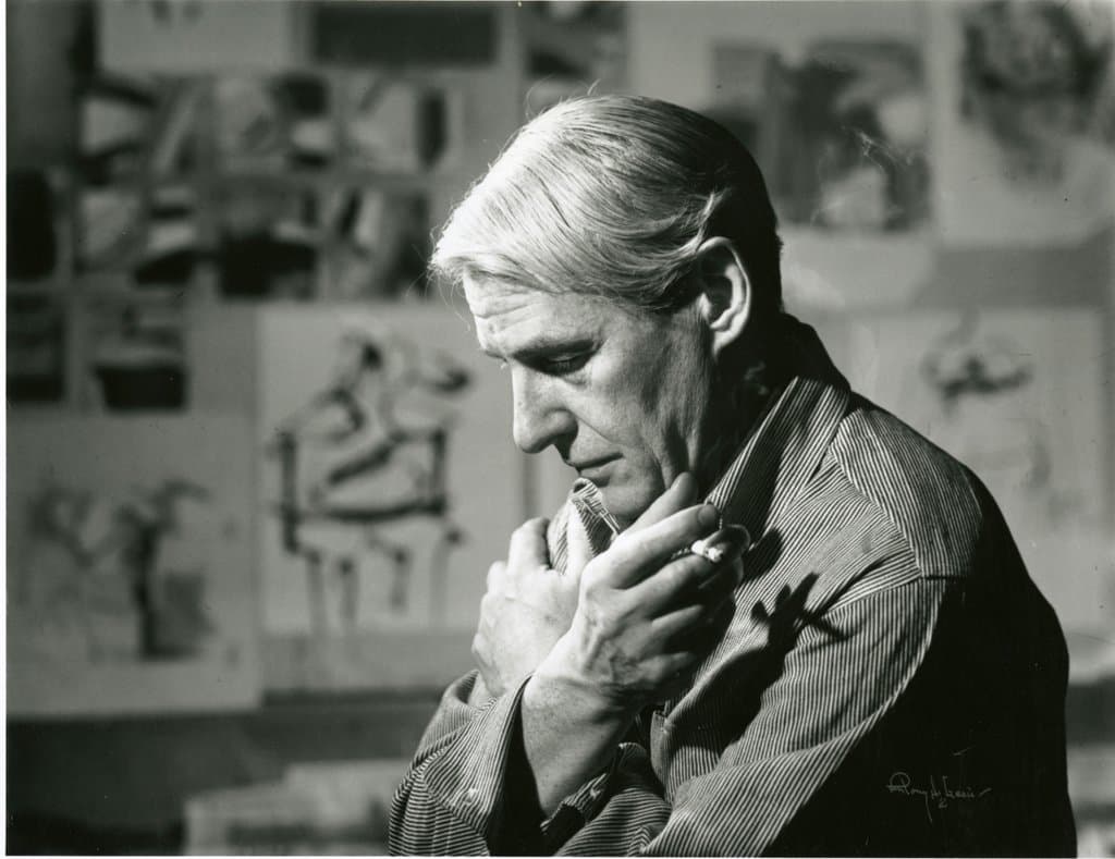 Willem de Kooning in his studio, hand to his chin, with drawings pinned on the wall behind.