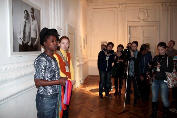 Zanele Muholi in a patterned shirt and black hat, with others in a gallery hung with portraits.