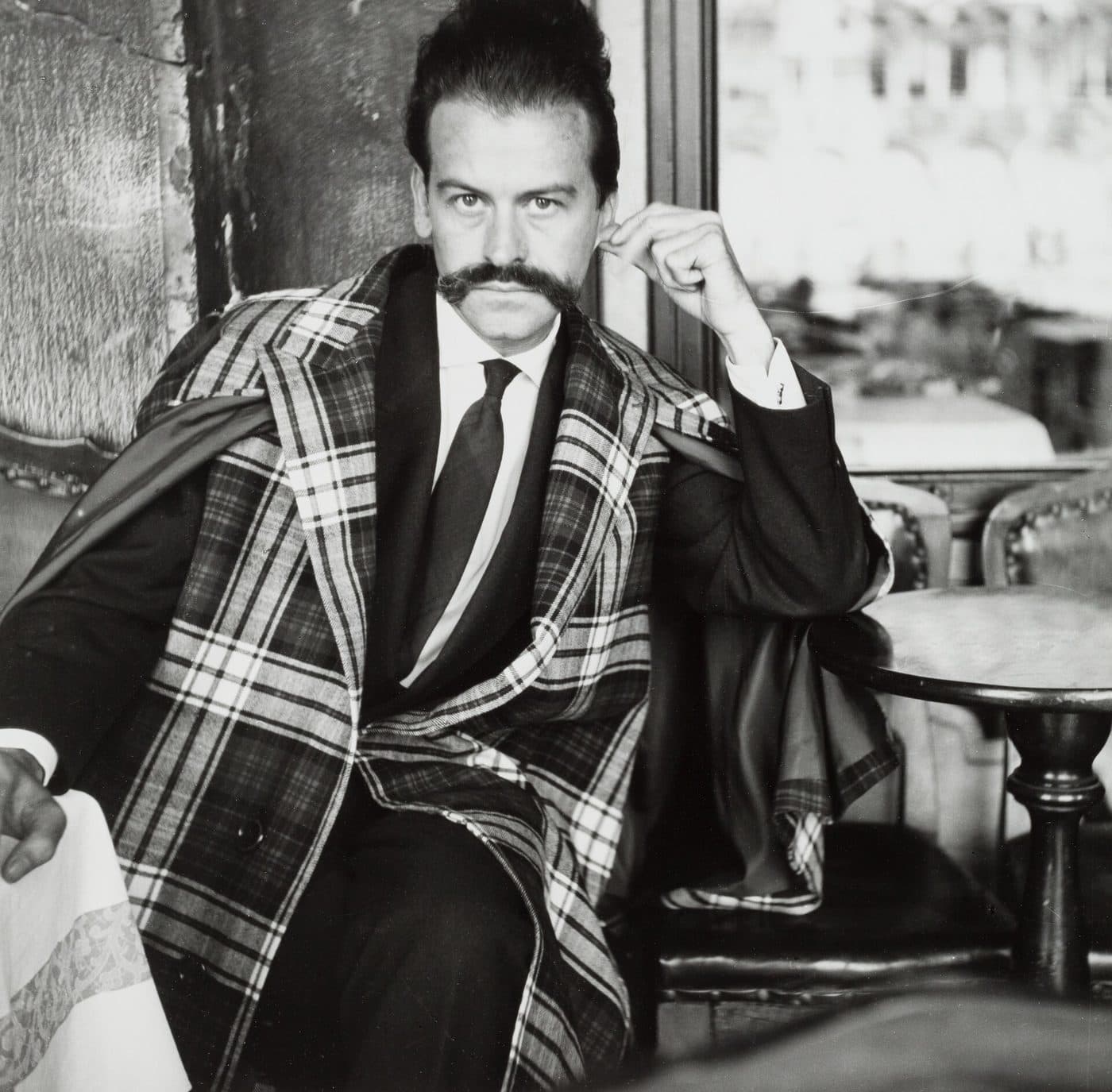 Georges Mathieu, with a long waxed moustache and a large checked coat over his suit, seated at a café table in 1961.