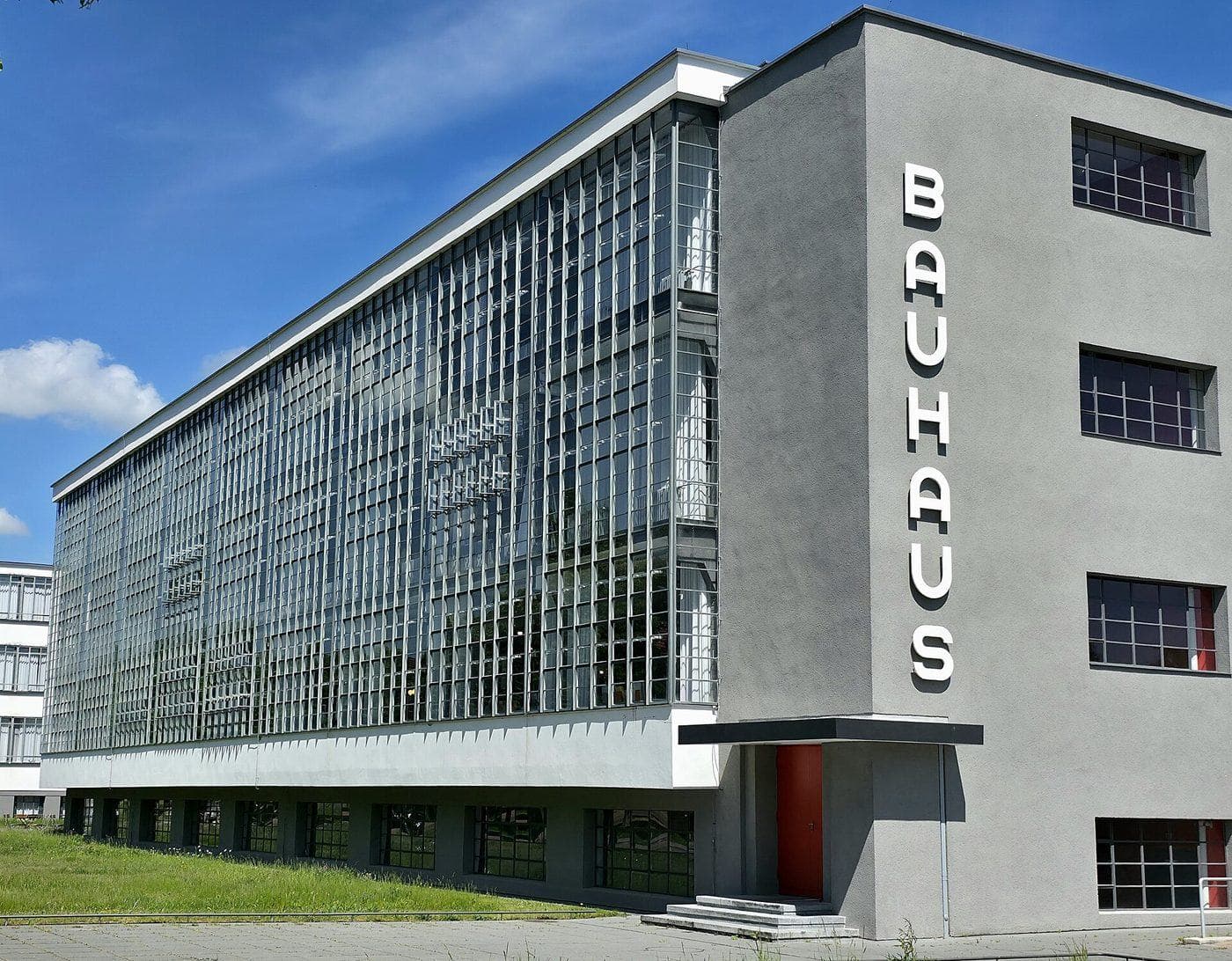 The Bauhaus building in Dessau: a grey block with a wall of glass and the word BAUHAUS running down its corner in white letters, under a blue sky.