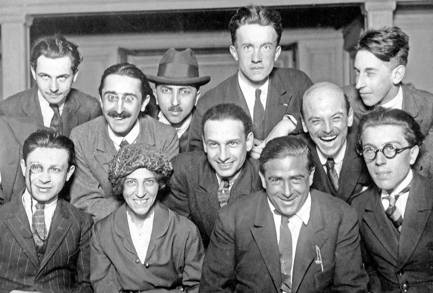 A group photograph of the Paris Dada circle in 1921, men in suits and one woman crowded together and smiling, Tristan Tzara with a monocle in the front row.