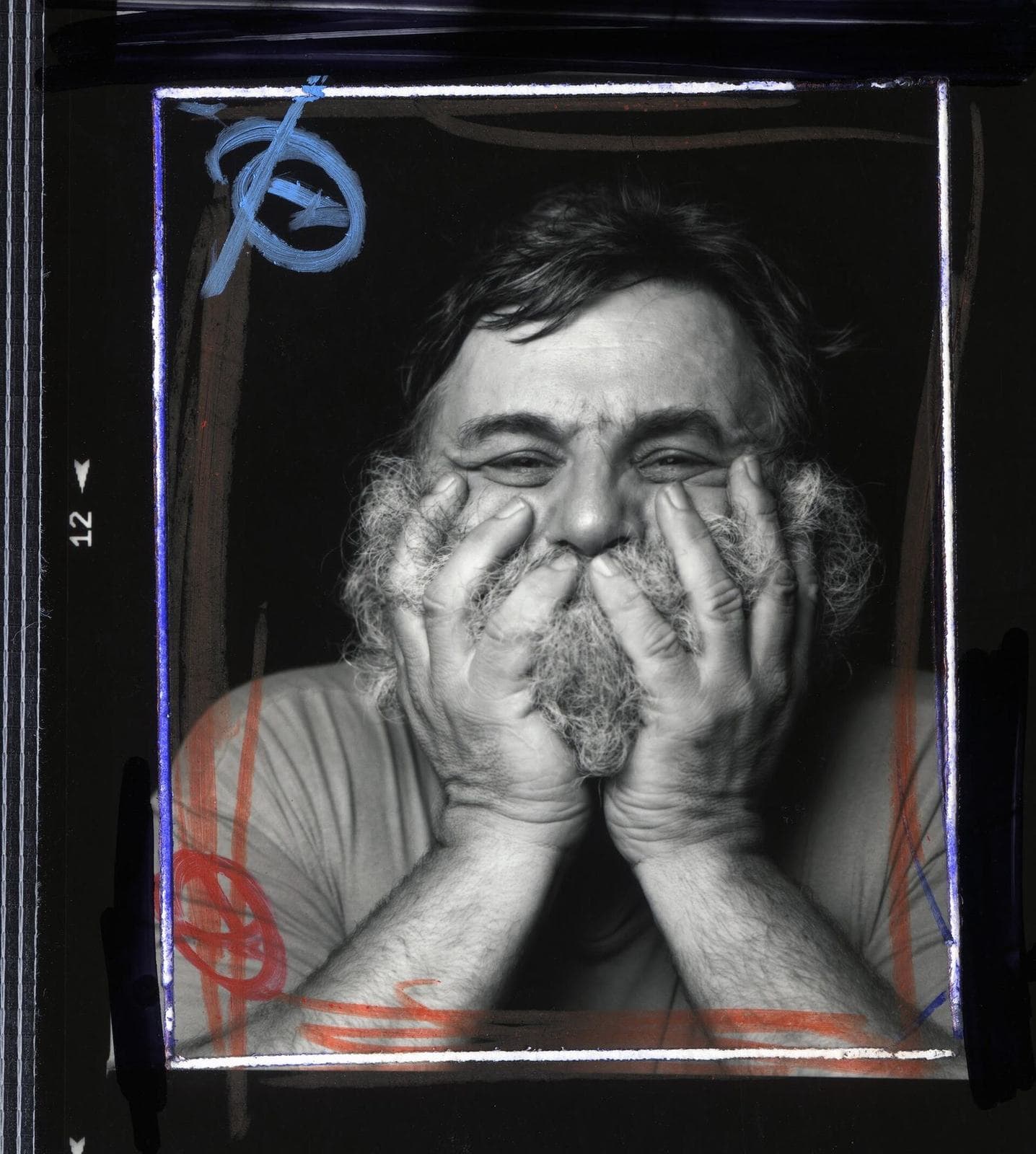 A. R. Penck grinning, his hands pressed to his bearded face, in a black-and-white photograph marked up in coloured crayon.