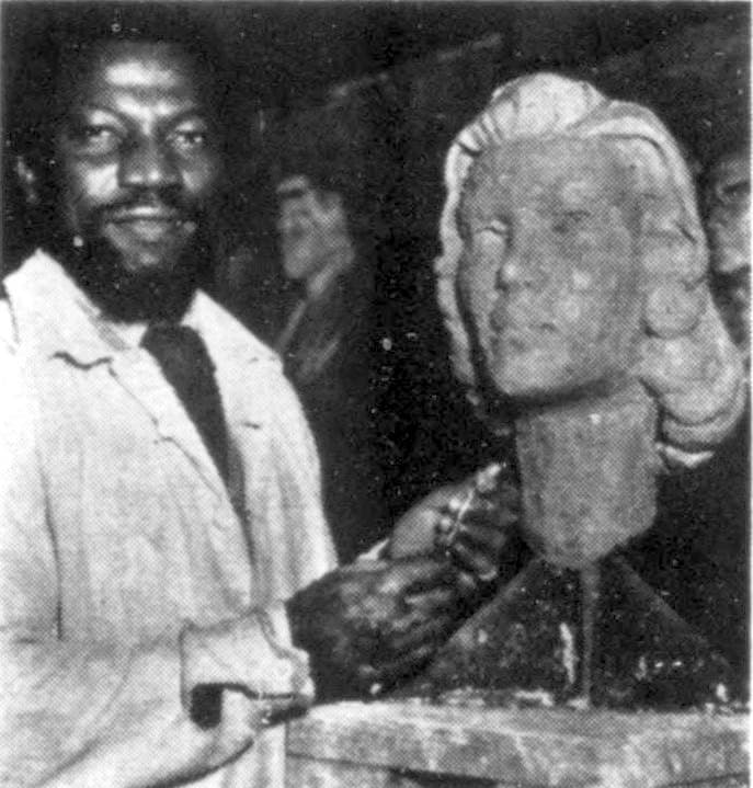 Ben Enwonwu in a work coat, smiling, beside the clay portrait bust of a woman that he is modelling, in a black-and-white photograph of 1957.